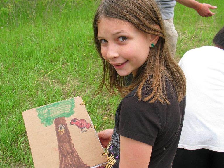 A girl stands in the grass with a drawing of a tree