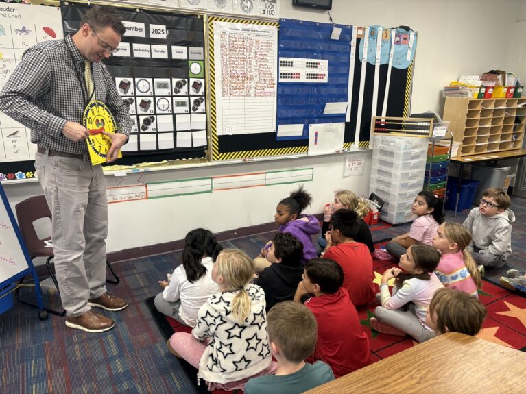 A teacher stands in front of a class of elementary students and teaches them how to tell time.