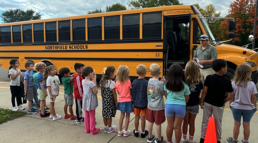Students at Spring Creek stand in a line facing a school bus, while the bus driver explains bus safety.