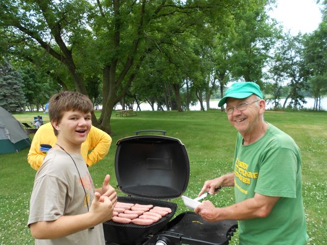 An older gentleman and a young boy stand smiling at the camera as they grill a multitude of hotdogs on a large grassy swath of land along the banks of the Canon river.